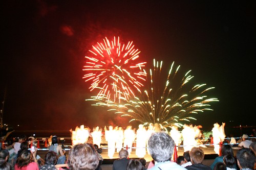 夜空に大きく鮮やかな赤と緑の打ち上げ花火が咲き乱れる中、観客の頭越しに舞台上でパフォーマンスを行っている人たちを捉えた写真