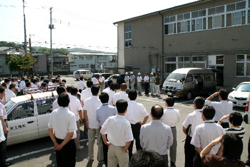駐車場で、大勢の関係者が、作業服姿の数人の職員などを囲んで、出発式を行っている写真