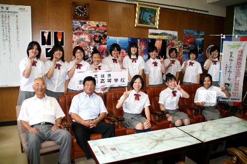 制服姿の女子高生たちが、座っている関係者の男性とともに、カメラに向かって笑顔でポーズを取っている集合写真