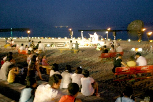 夜の砂浜に設置されたベンチに座った観客たちが、遠くの特設ステージで繰り広げられる夜のイベントを鑑賞している様子の写真