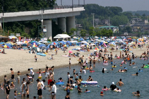 青空が広がる晴れた日に、大勢の海水浴客が海に入っており、色とりどりのパラソルやテントが並ぶ賑やかな海水浴場の写真