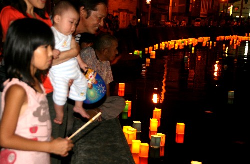 夜の川辺で、家族連れなどの人々が水面に浮かぶ無数の温かい光を放つ灯篭流しを真剣に見つめている厳かで美しい伝統行事の写真