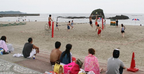 海岸沿いで、数人がビーチバレーを行っており手前の段差のある舗装エリアの場所に座った観客たちがその様子を見守っている写真