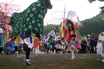 屋外の広場で、緑色の布地に白い花の模様が入った獅子舞と、笠をかぶり鮮やかな衣装を着た演者が、大勢の観客を背景に対峙して舞を披露している様子の写真