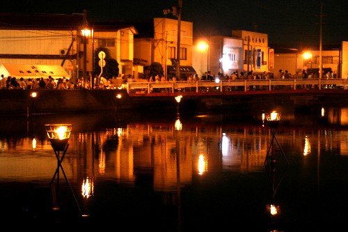 暗い夜の水面に、建物群の温かい光が反射し、手前に置かれた3本の脚に支えられたかがりびの灯りが厳かに揺らめく、情緒的なかがり火祭りの夜景写真