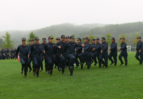 制服と帽子を着用した大勢の消防団員が、芝生の広場の上を力強く行進している訓練の様子の写真