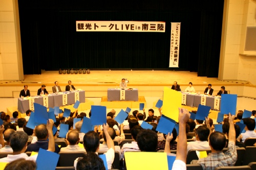 「観光トークLIVE in 南三陸」の横断幕が掲げられた会場で、聴衆が青や黄色のボードを高く掲げており、壇上のパネリストたちと意見交換をしている講演会の写真