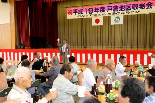 「祝 平成19年度 戸倉地区敬老会」と書かれた横断幕と日の丸の下、高齢者たちがテーブルを囲み、ステージ上の男性の音頭で一斉にコップを掲げて乾杯をしている敬老会の写真