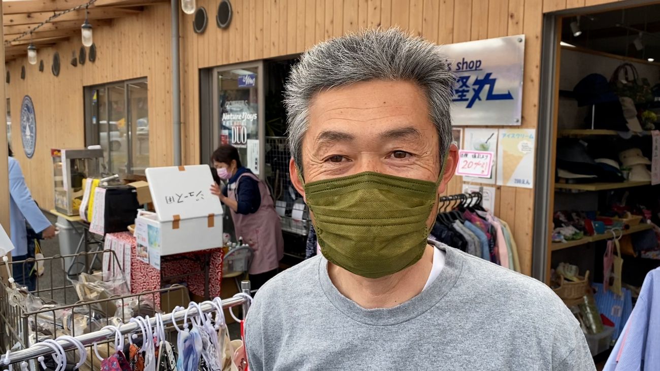 ハンガーラックにかけられた服が並んだ店や、隣の店先でエプロンを着た女性がジュース用と手書きで書かれた発泡スチロールの箱の蓋を開けている長屋の様な木造の建物の前に立つ、緑色のマスクをした牧野会長がカメラを見ている写真