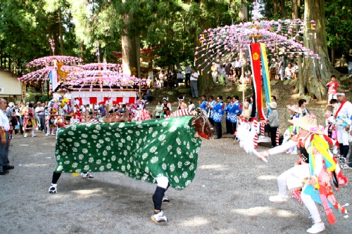 神社の境内で、緑色の胴体と黒い頭の獅子舞が、鮮やかな衣装を身につけた演者と対峙し、飾り付けがされた豪華な山車や大勢の観客を背景に舞っている村祭りの写真