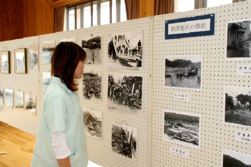女性が、壁一面に展示された「新湊地区の惨状」と題された被災地のモノクロ写真を見つめている様子を捉えた写真