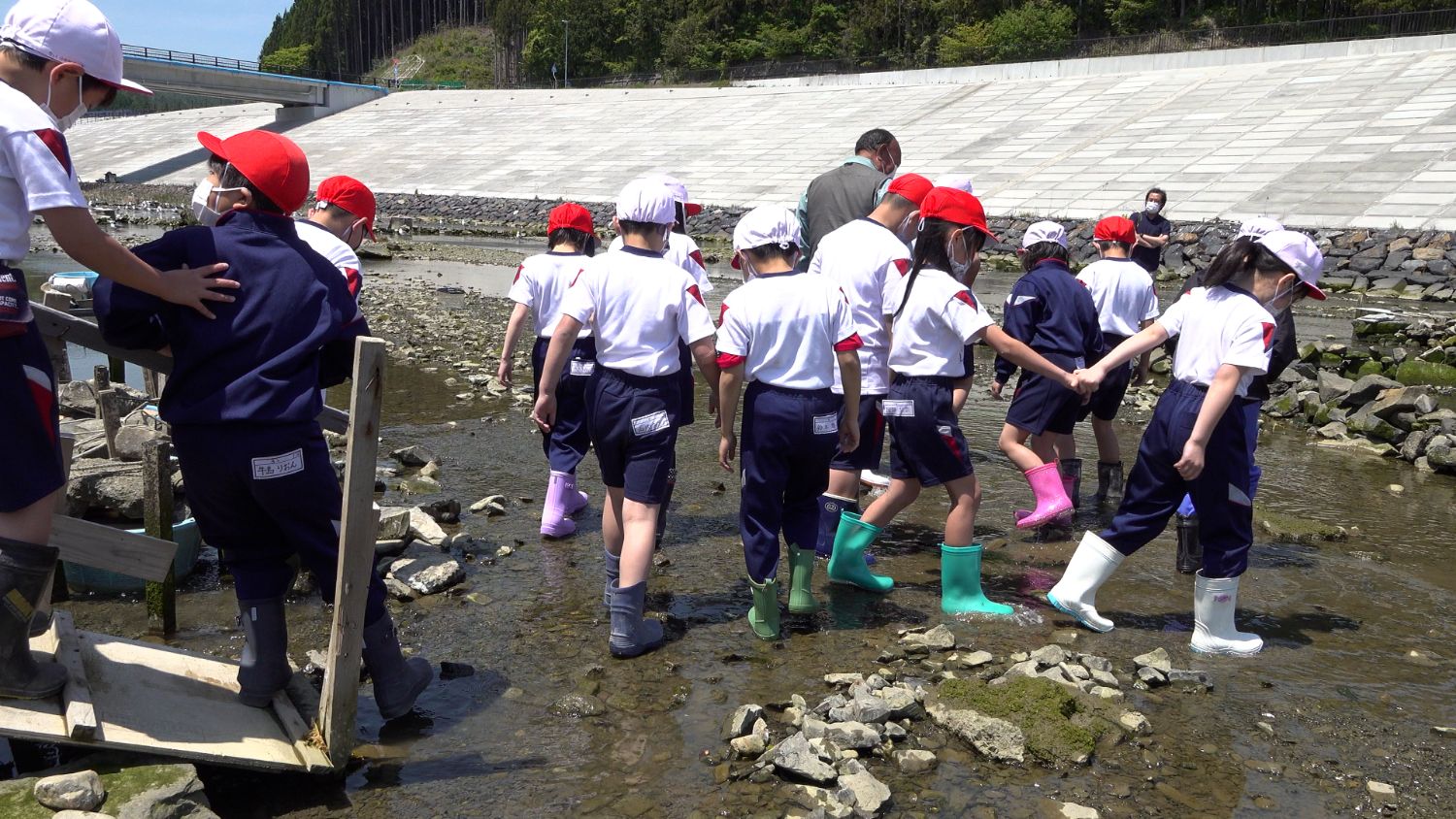 児童たちが長靴を履いて川の中を歩き、川の生き物や環境を調べる体験学習に参加している写真