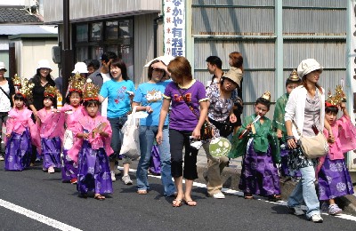 屋外の舗装された通りで、ピンク色と紫色の鮮やかな衣装を身につけ、金色の冠をかぶった複数の幼い子どもたちが、引率の大人たちと共に、行列に参加している様子の写真