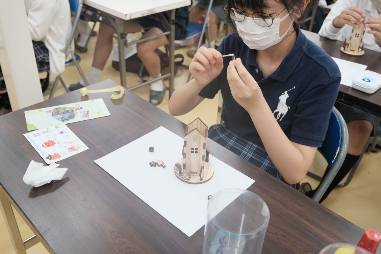 教室で青い服を着た児童が木製のペンスタンド型模型のパーツを手に取り組み立て作業を行っている様子の写真