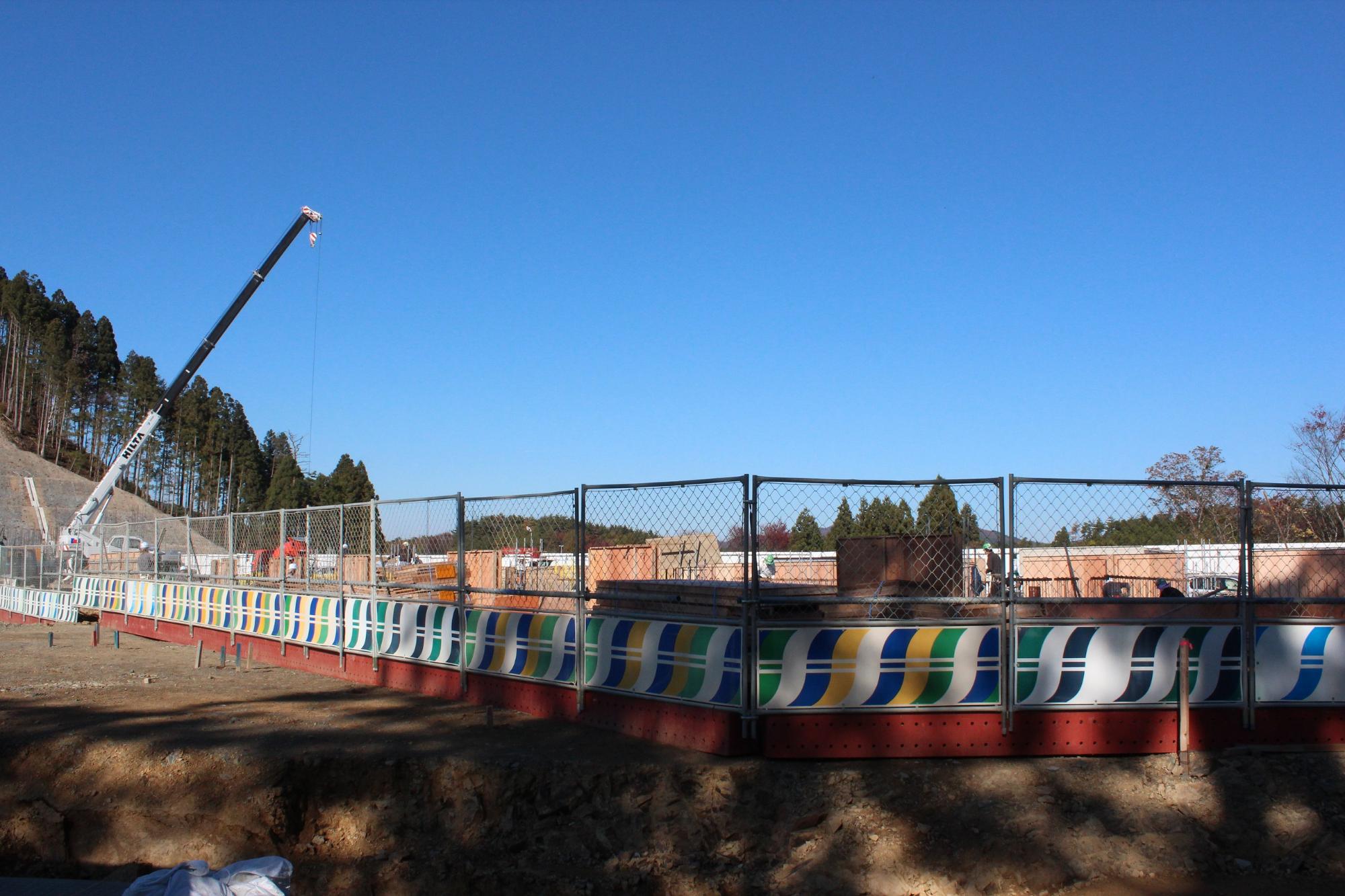 快晴の青空の下、色鮮やかな防護柵に囲まれた建設現場では、クレーン車が資材を吊り上げており、奥には整備中の基礎構造や作業員、背後に木々と丘が見える写真