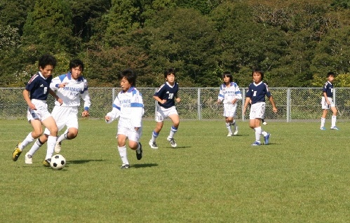 芝生のグラウンドで、白いユニフォームと紺色のユニフォームを着た子どもたちが、サッカーの試合に熱中している写真