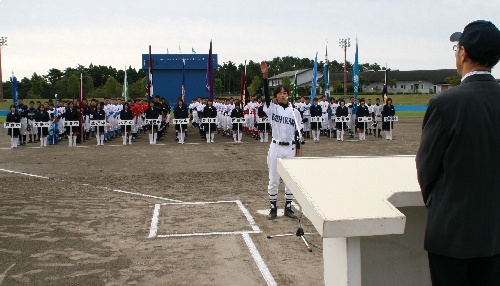 大勢の高校球児が整列した野球場のグラウンドで、ホームベース前に立つユニフォーム姿の代表生徒が、右手を挙げて宣誓をしている大会の写真