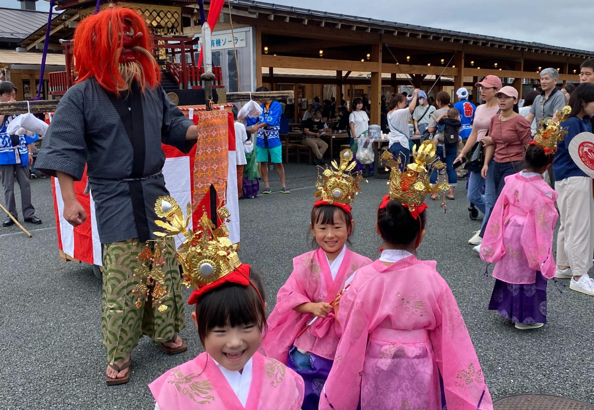 華やかな冠をかぶった女の子たちが笑顔で歩き、赤い仮面をつけた大人や見物客で賑わっている写真