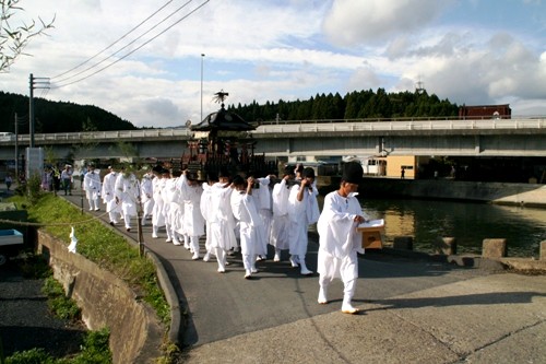 川沿いの道を、白い装束と黒い烏帽子をまとった関係者が、木造の豪華な神輿を伴い、一列になって歩いている秋祭りの様子の写真