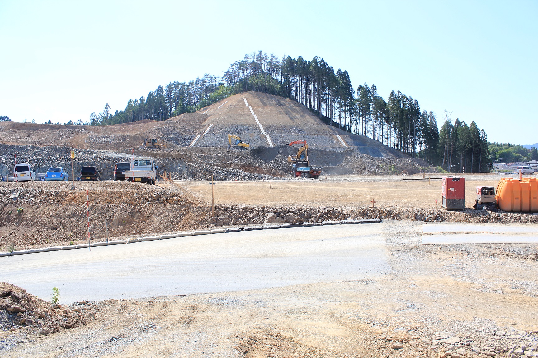 山のふもとで大規模な造成工事が行われており、階段状に整地された斜面には複数のショベルカーが作業を進め、手前には舗装された道路と工事車両、資材が並んでいる様子が広がる写真