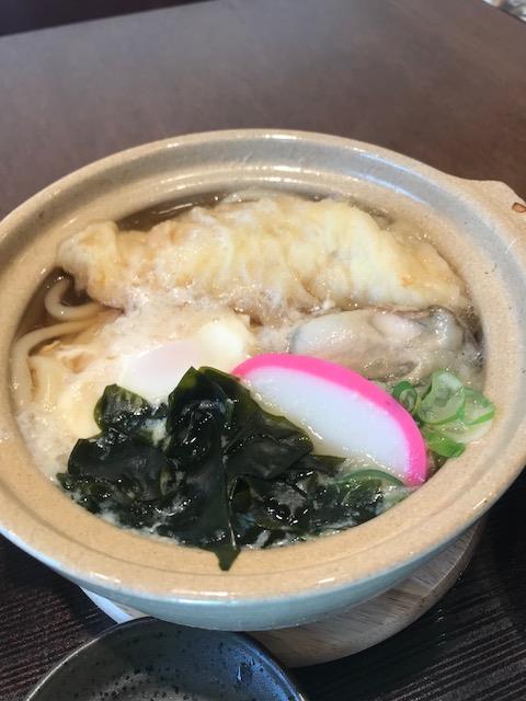 土鍋の中に、うどん、牡蠣、かまぼこ、わかめ、ねぎ、天ぷら、卵などが入った熱々の鍋焼きうどんがテーブルの上に置かれている写真