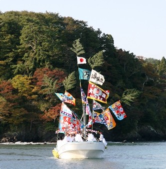 穏やかな海上で、大漁旗や日の丸をたくさん立てた白い小型船が、紅葉が始まった緑の山を背景にして航行している写真