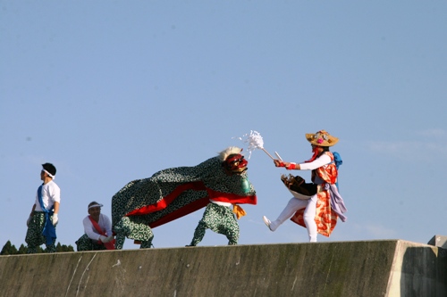 青空を背景に、コンクリートの上で、獅子舞と、両手に白い棒を持った演者が向かい合って踊りを披露している、伝統的なお祭りの写真