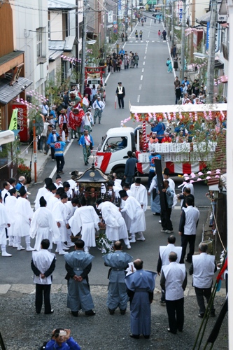 両脇に家々が並ぶ坂道で、白い装束の担ぎ手たちが神輿を担いでおり、その後ろには飾り付けられたトラック山車や多くの見物客が見える祭りの写真