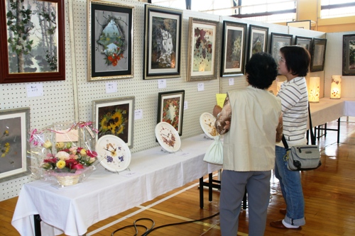 広い会場で、二人の女性が、壁に掛けられた作品とテーブルに並べられた花や絵皿などの展示物を鑑賞している様子の写真