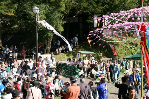 快晴の屋外の広場で、大勢の観客が見守る中、獅子舞の演舞が披露されている秋祭りの写真