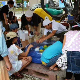 屋外の祭り会場で、色とりどりの風船が浮かぶ小さなビニールプールを囲んで子供たちが遊ぶ様子を写した縁日の様子の写真