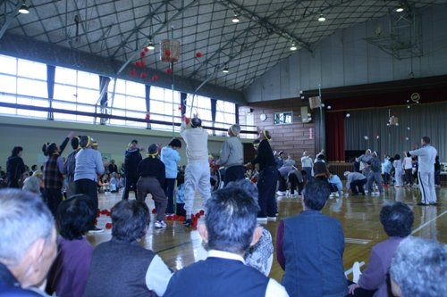 体育館で、黄色いハチマキを締めた人々が、籠に向かって赤い玉を投げ入れている玉入れ競技のようなレクレーション行っているイベントの様子の写真