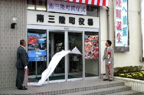 「南三陸町開庁式」の看板と「南三陸町役場」の文字が掲げられた建物の正面玄関で、スーツ姿の二人の男性が、白い布と紅白の紐を使った除幕式を行っている様子の写真