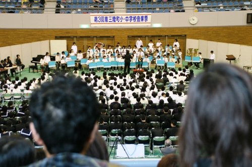 「第3回 南三陸町小・中学校音楽祭」と書かれた看板の下、体育館のフロアに集まった大勢の小中学生が聴衆として座り、前方のステージで吹奏楽団が演奏を披露している写真