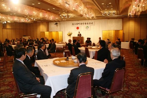 華やかなシャンデリアが輝くホテルの宴会場のような場所で、「南三陸町功労者表彰式」と書かれた看板の下、スーツ姿の多くの参加者がテーブルに着席し、中央の演台に立つ人物の進行を見守っている式典の写真