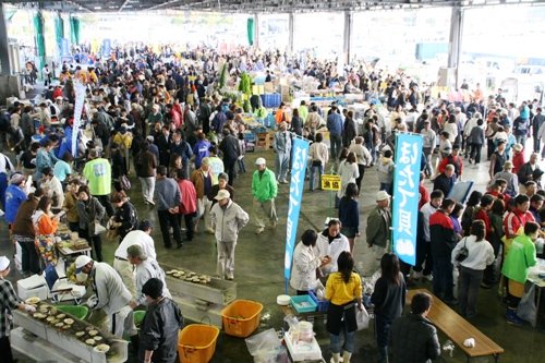 市場のような広い屋内に、老若男女の大勢の人々が集まり、中央の通路を進んだり、食べ物や商品を売る屋台のようなブースに立ち寄ったりしている、賑やかなイベント会場の写真