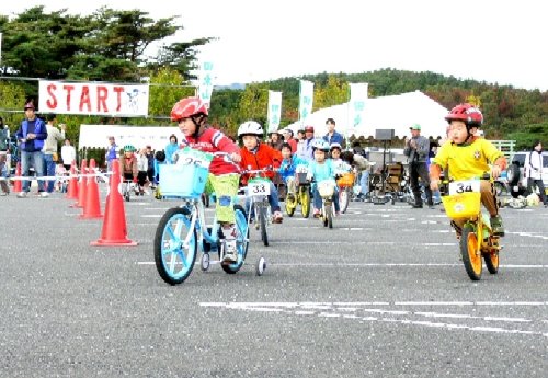 ヘルメットを着用した複数の小さな子どもたちが、補助輪付きの自転車などに乗って、「START」と書かれた看板の前から一斉にスタートしているイベントの様子の写真