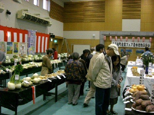 広い屋内の会場で、多くの来場者が新鮮な野菜や特産品が並べられた展示テーブルを見て回っている、「産業フェア」歌津会場の様子の写真