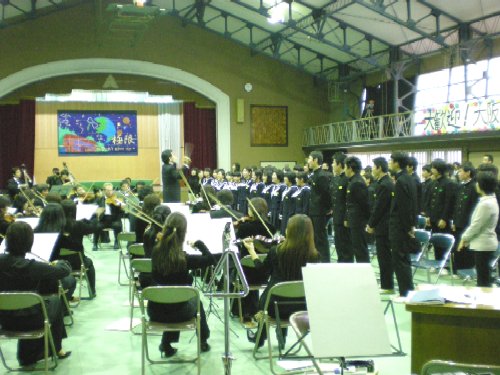 体育館で、指揮者を中心にオーケストラが演奏し、制服を着た大勢の生徒が立って合唱している様子の写真