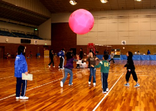 広々とした体育館で、数人の子供たちが、関係者に見守られながら、天井近くに打ち上げられた巨大なピンク色のボールを使ってスポーツを楽しんでいる様子の写真