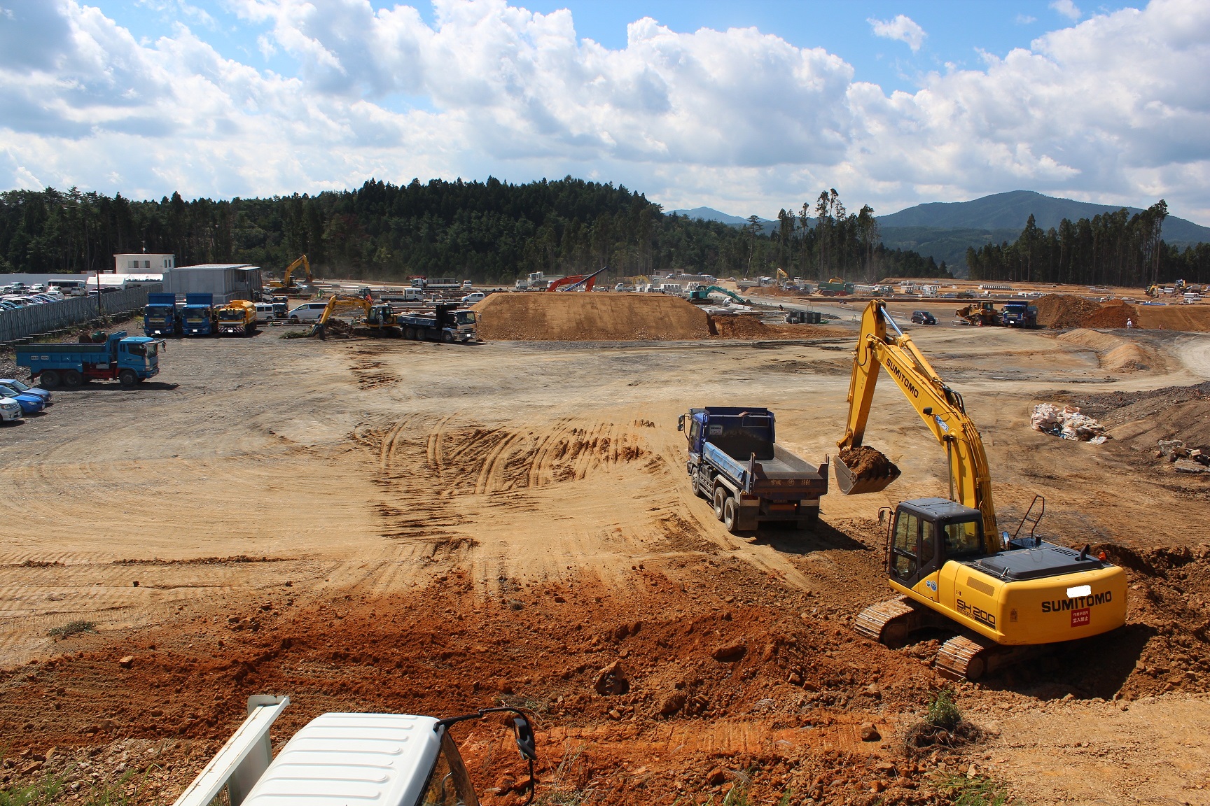 青空の下、広大な造成地で多数の重機やダンプトラックが稼働し、手前では黄色いショベルカーが土を積んでトラックに積載しようとしている写真