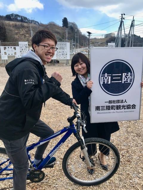 広場で青い自転車にまたがった佐藤さんと、一般社団法人南三陸町観光協会の看板を両手で掲げた女性リポーターが笑顔で並んでいる写真