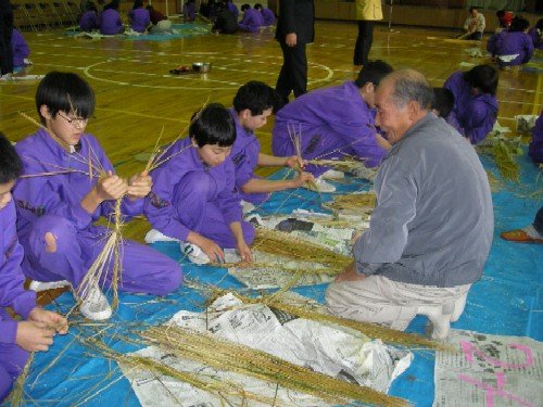 体育館の床に敷かれた青いシートの上で、紫色のジャージを着た生徒たちが藁を使い関係者の男性から手ほどきを受けながらしめ縄を制作している写真