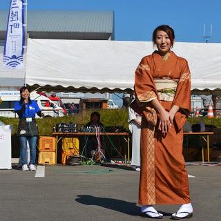 音響機材が並んでいる白いテントの下で青色のシャツを着た女性が笑顔で進行しているなか、ベージュ色の帯に橙色の着物を着た女性が屋外イベント会場で笑顔で立っている写真