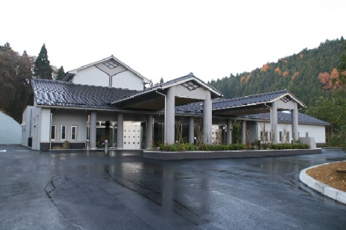 落ち着いた灰色と白を基調とし、黒い瓦屋根が特徴的な建物で、エントランス部分が大きな庇と柱で覆われている斎場施設の外観写真