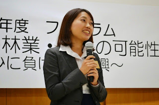 背後に横断幕が掲げられている会場の前に立つスーツ姿の女性が、マイクを持って話している様子をアップで撮影した写真