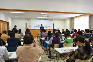 会議室のような会場で多くの参加者が前を向いて着席し、正面のスクリーン横では男性が話している写真