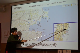 南三陸町の地図が映し出されたスクリーンの前に立つ男性が、長い指示棒で地図を指しながら説明している写真