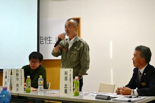 名前の表示札や飲み物が置かれている長机の席の登壇者3名のうち、中央の男性がマイクを持って発言している写真