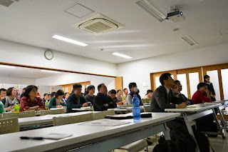 長机に座った参加者たちが真剣な表情で講演を聞いている様子を、左前方から撮影した写真